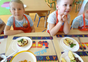 Uśmiechnięte dzieci siedzą przy stole, przed sobą mają talerze na których są przygotowane przekąski z wafli ryżowych.